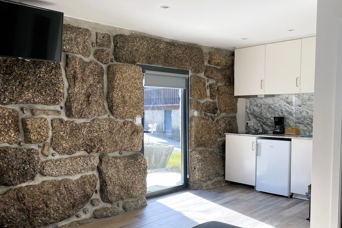 Una habitación moderna con paredes de piedra, un televisor de pantalla plana, una cama con toallas, una pequeña cocina con cafetera y vistas a través de una puerta de cristal.