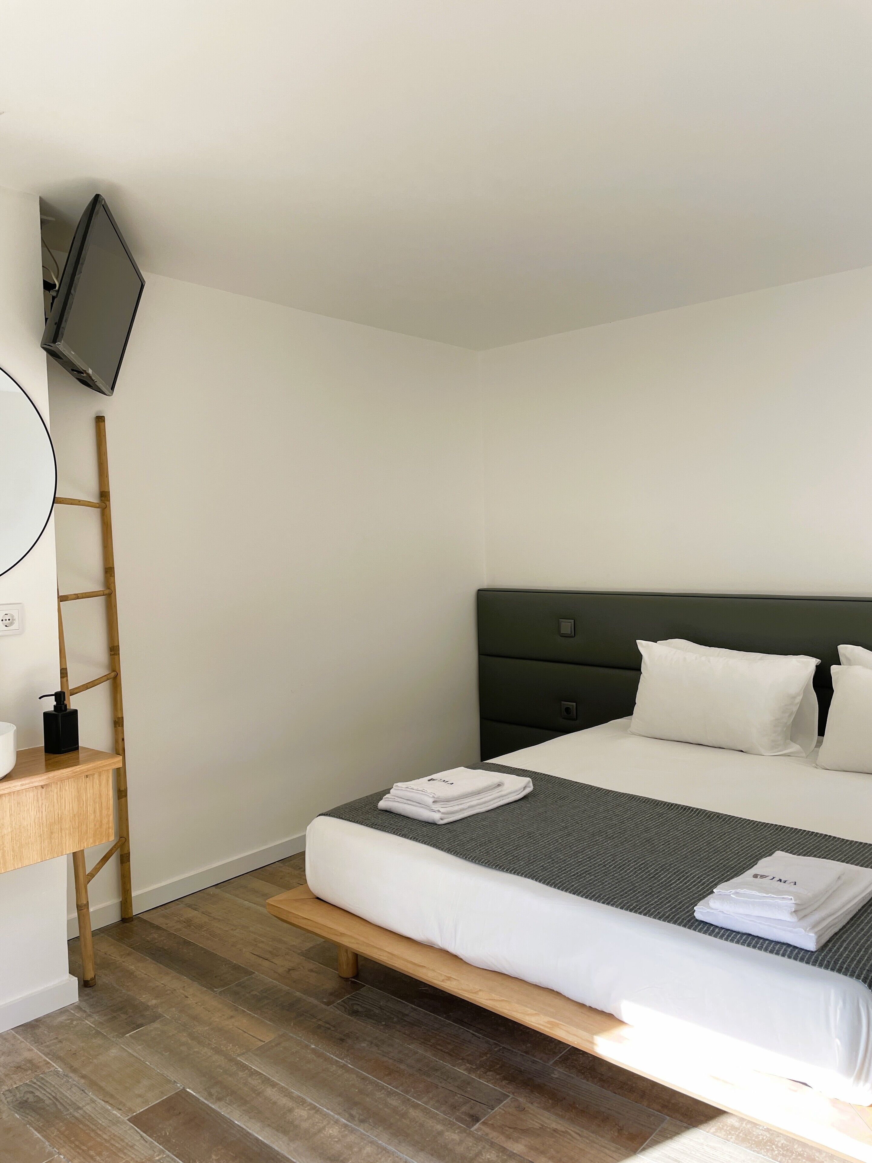 A neatly made bed with white linens, a wooden bedside table with a black bottle, and a television mounted on the wall.