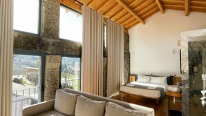 Una habitación de hotel moderna con techo de madera, un ventanal con cortinas, un sofá, una cama y un baño con lavamanos de mármol.