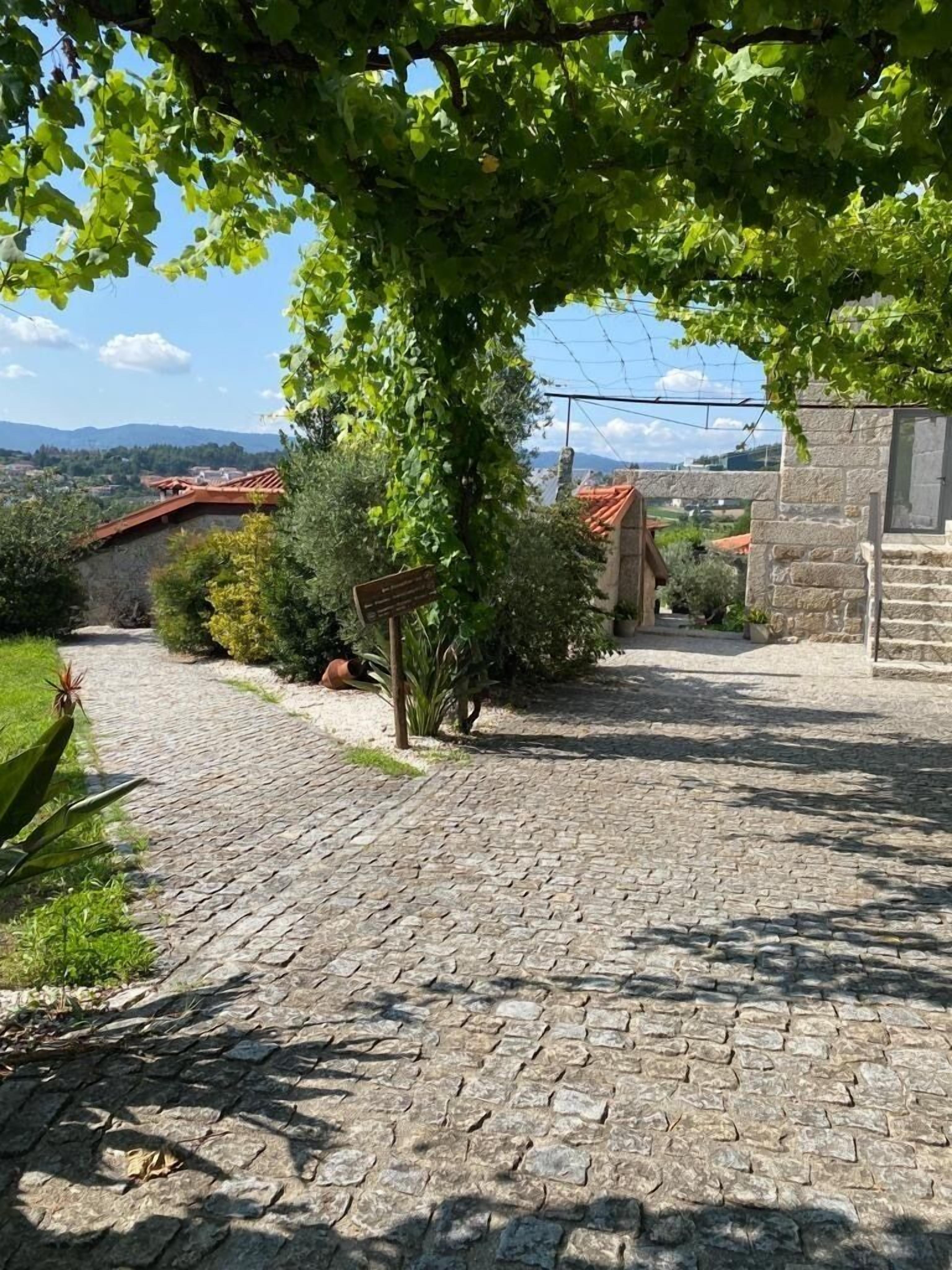 A cobblestone pathway leading to a stone building with a red-tiled roof, surrounded by greenery and a clear view of distant mountains.