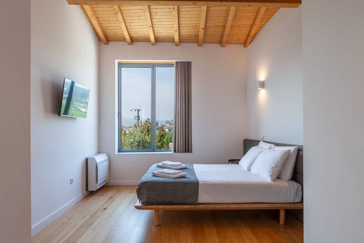 Un dormitorio con una cama, un televisor montado en la pared, un ventanal con vistas y un techo con vigas de madera.