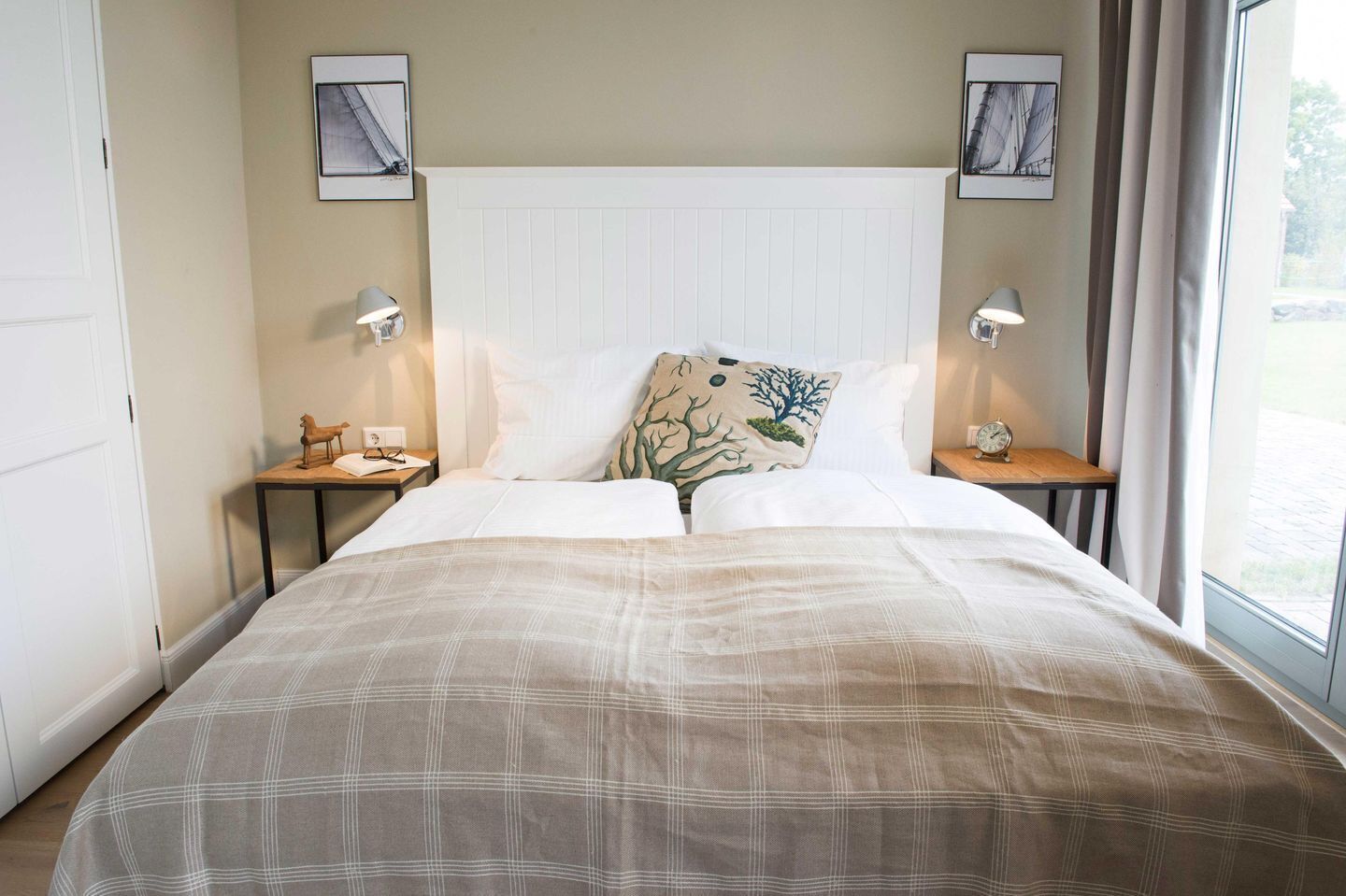 A neatly made bed with a plaid bedspread, two bedside tables with lamps, a wall-mounted clock, and two framed pictures above the bed.