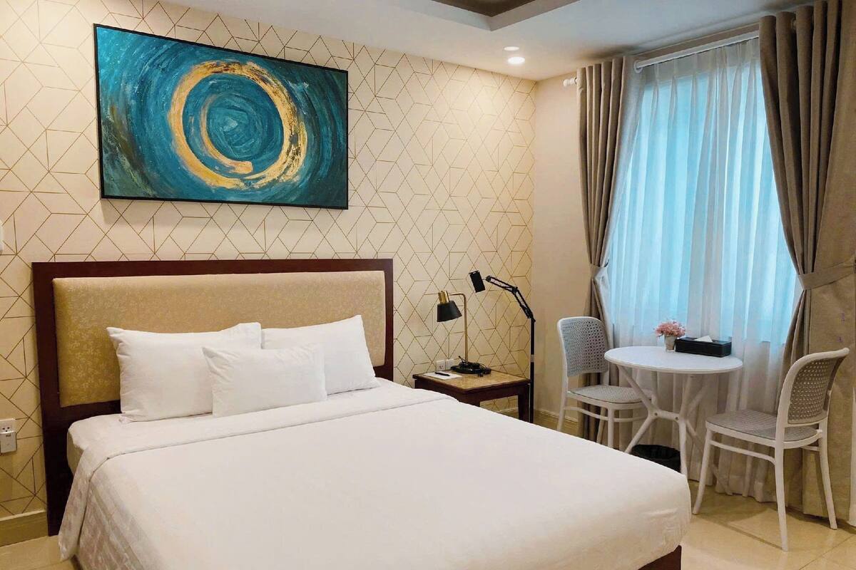Quarto de hotel com cama, escrivaninha, cadeira, luminária e um quadro na parede.