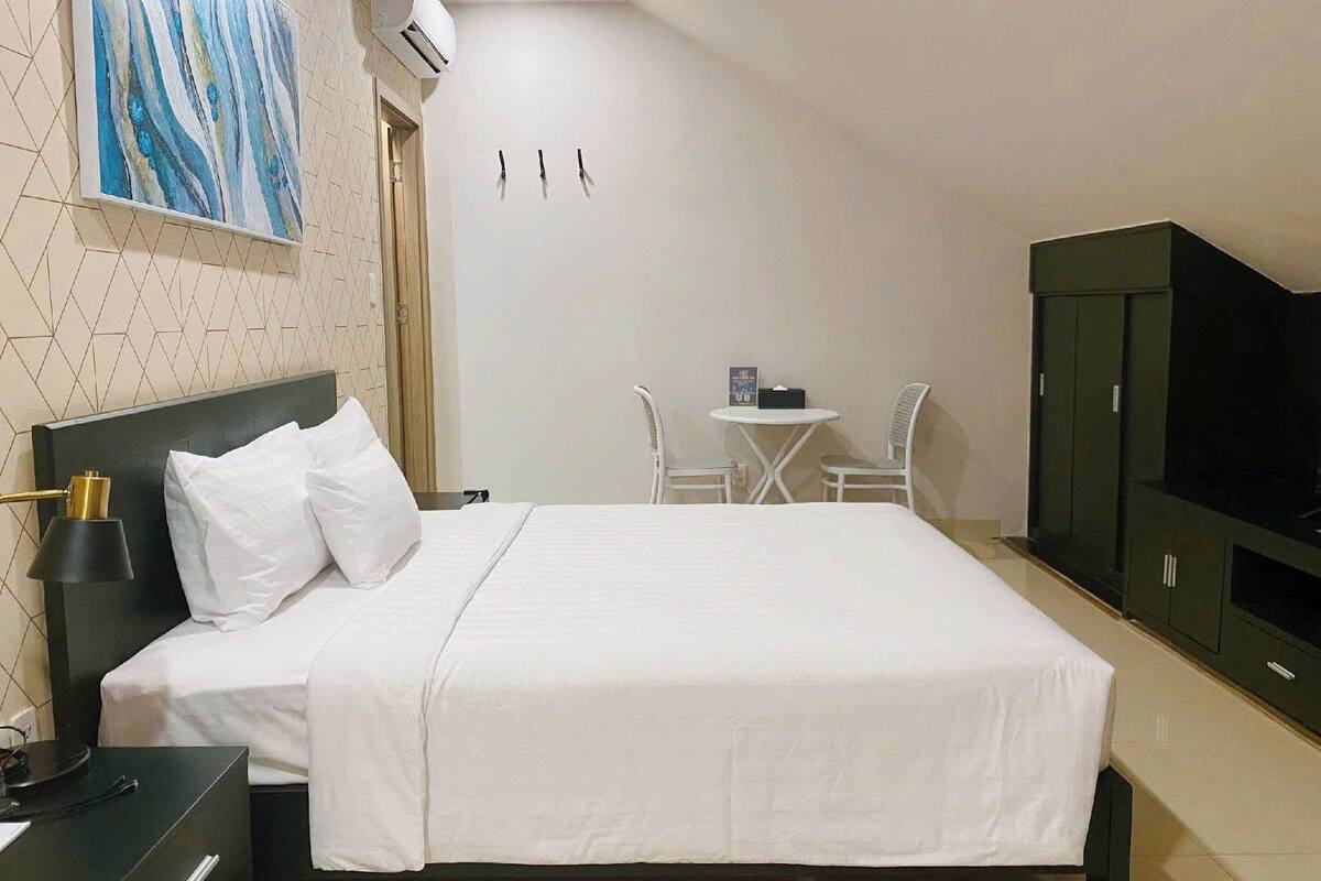 Quarto de hotel com cama, mesa de cabeceira, luminária, televisão, guarda-roupa e uma pequena mesa com cadeiras.
