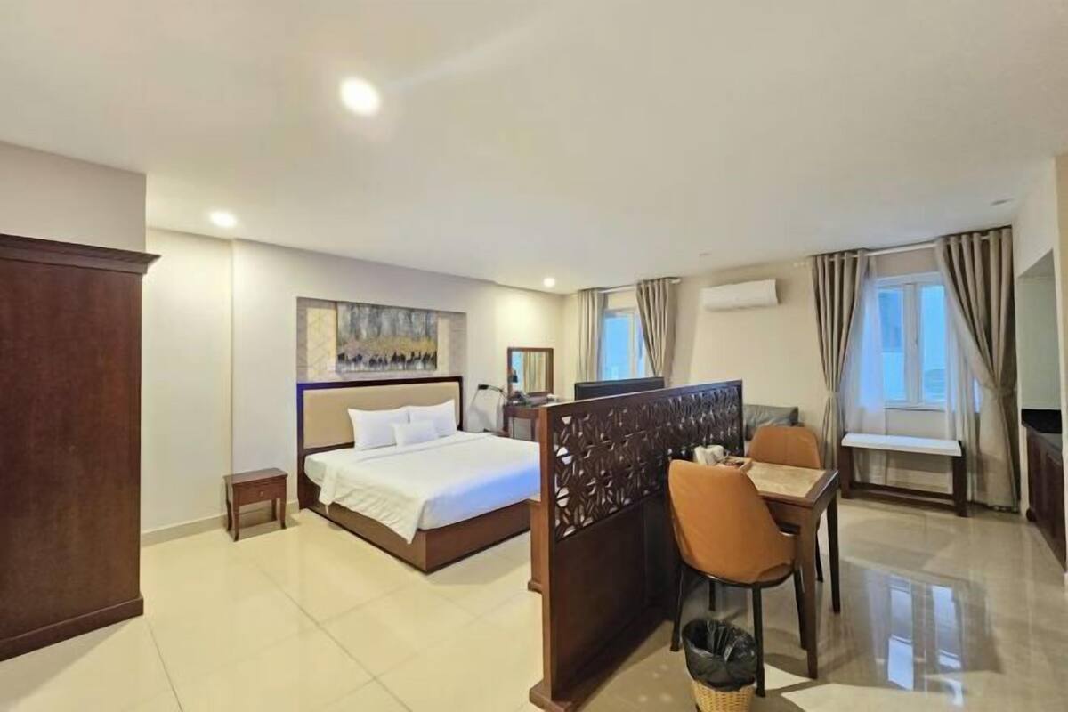 Quarto de hotel com cama, escrivaninha, cadeira, mesinha, armário e janela com cortinas.