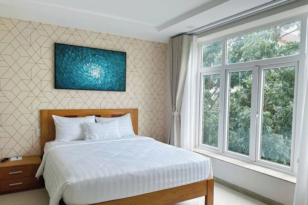 Uma cama bem arrumada com lençóis brancos, cabeceira de madeira e uma janela grande com cortinas.