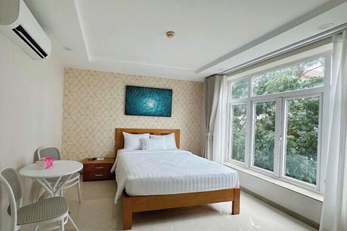Een slaapkamer met een bed, een klein tafeltje en een stoel. Er is een raam met uitzicht op groen.