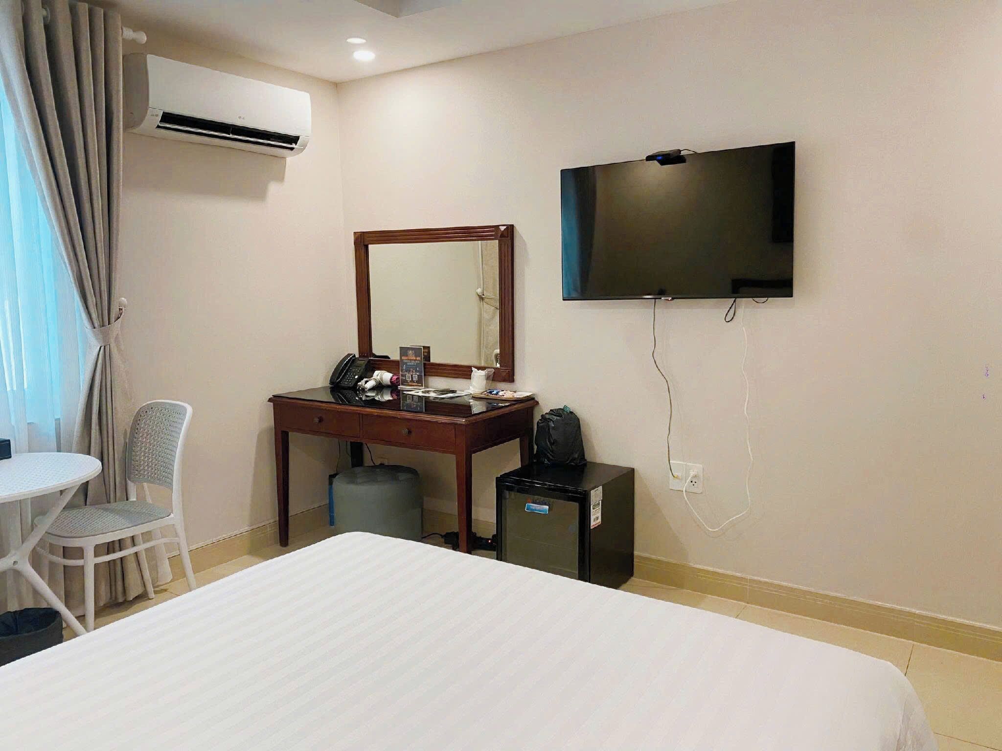 A hotel room with a bed, a desk with a telephone, a small refrigerator, a television mounted on the wall, and a window with curtains.
