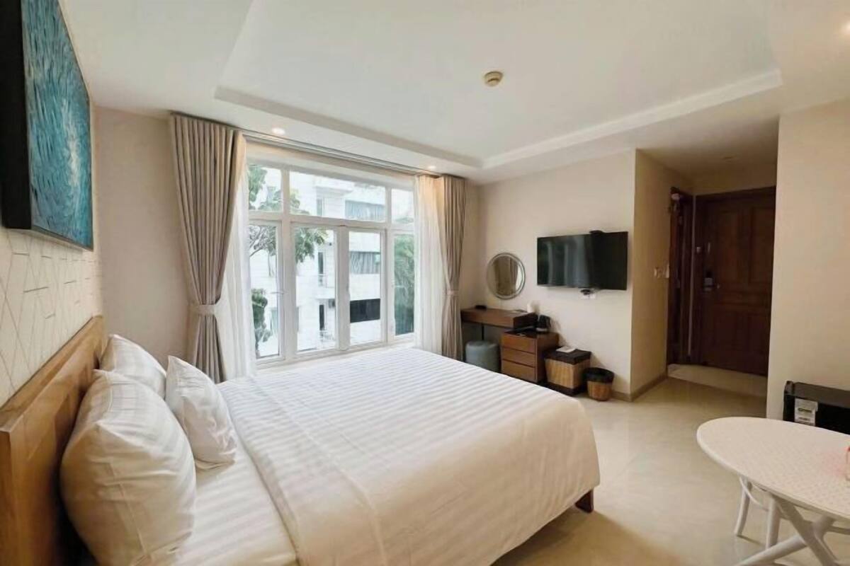 Quarto de hotel com uma cama grande, escrivaninha, cadeira, televisão e janela com cortinas.