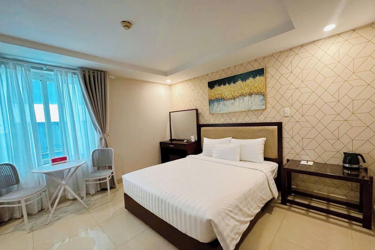 Quarto de hotel com cama, escrivaninha, cadeira, janela com cortinas e um quadro na parede.
