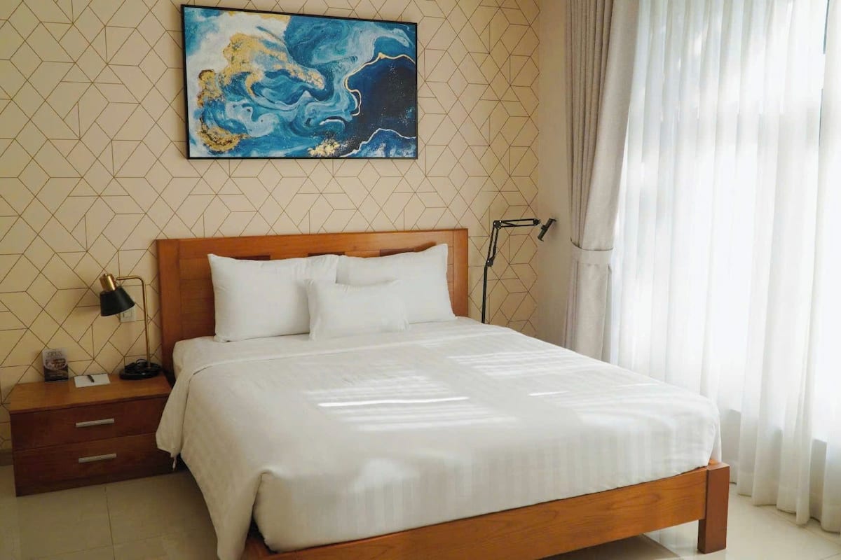 Quarto de hotel com cama de madeira, roupa de cama branca, mesa de cabeceira, luminária, quadro na parede e cortinas leves.
