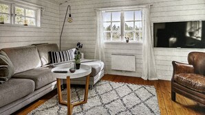 A cozy room with a wooden ceiling, a sofa, a coffee table, a TV, and a window with a view of greenery.