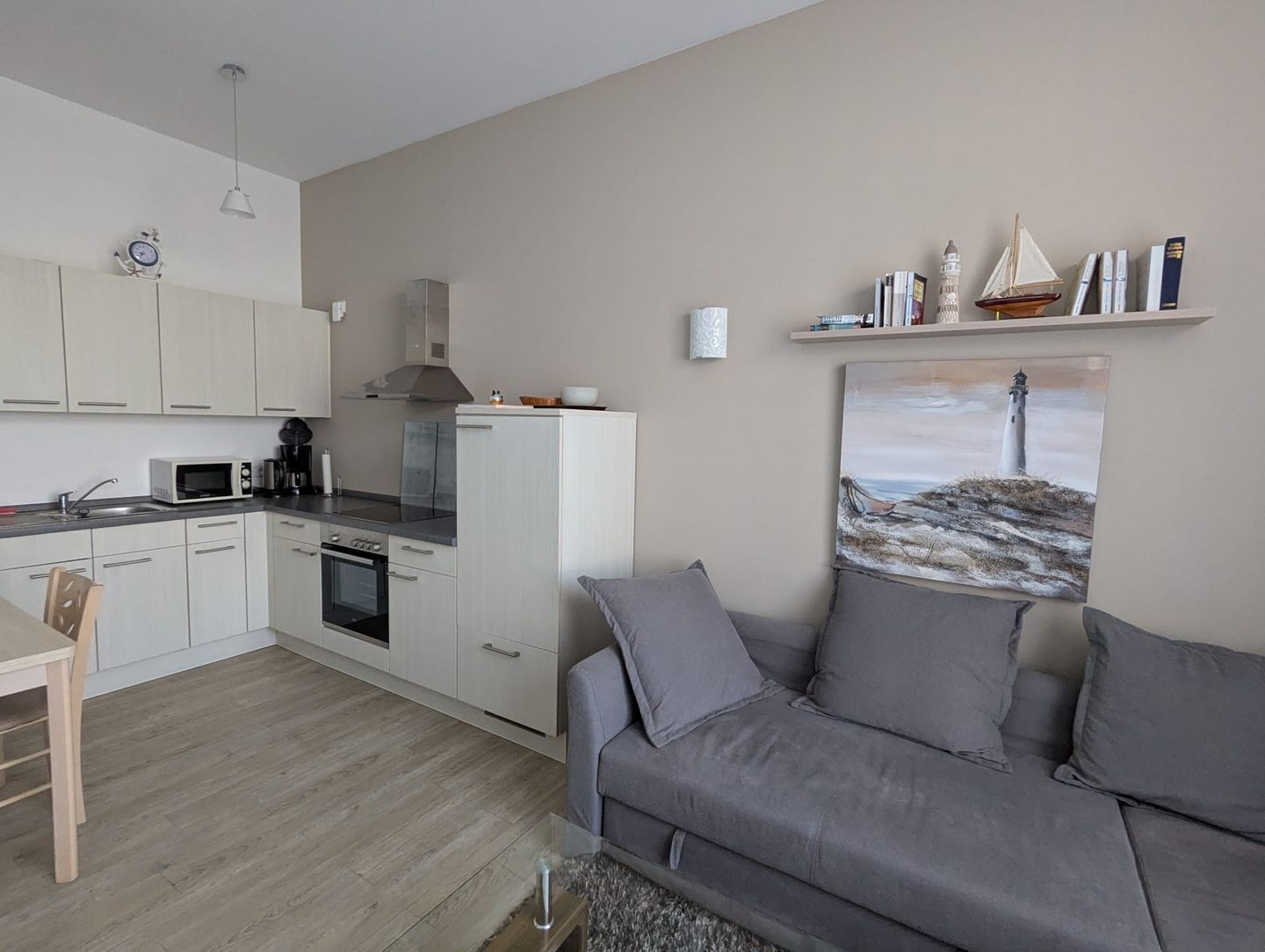 A modern kitchen with white cabinets, a microwave, and a stove. There's a gray sofa and a painting of a lighthouse on the wall.