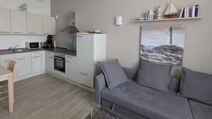 A modern kitchen with white cabinets, a microwave, and a stove. There's a gray sofa and a painting of a lighthouse on the wall. - Herold RE-01 - Cozy vacation apartment The Remise Herold on Usedom with (Stolpe auf Usedom)