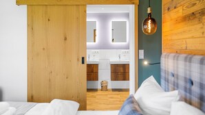 A bedroom with a wooden door, a bed with white bedding, a plaid headboard, and a view into a bathroom with a mirror and two sinks. - Studio Bairerkopf | Pool, sauna & mountain view (Reit im Winkl)