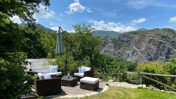 Un espace extérieur aménagé avec des meubles en osier et une table, entouré de verdure et avec une chaîne de montagnes en toile de fond.
