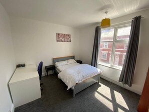 Un dormitorio con cama, escritorio, silla, ventana con cortinas y un cuadro en la pared.