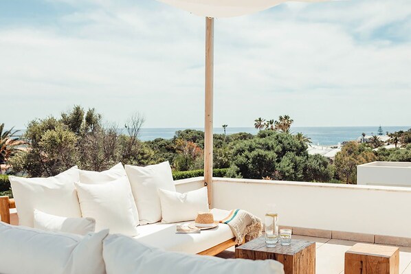 A balcony with a white sofa, a wooden table with glasses, and a view of the sea and greenery.