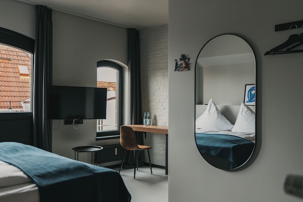 Quarto de hotel com cama, escrivaninha, cadeira, televisão e um espelho grande.