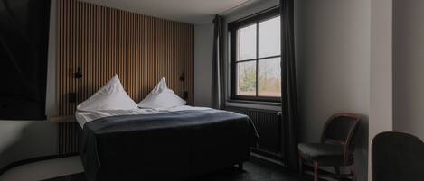 A modern hotel room with a large bed, a chair, a window with curtains, and a wall-mounted TV.