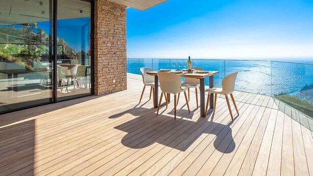 A modern outdoor dining area with a wooden deck, a glass wall, and a view of the ocean. - My Rental Homes | Villa Mirante  with sea view and private pool (Begur)