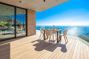 A modern outdoor dining area with a wooden deck, a glass wall, and a view of the ocean. - My Rental Homes | Villa Mirante  with sea view and private pool (Begur)