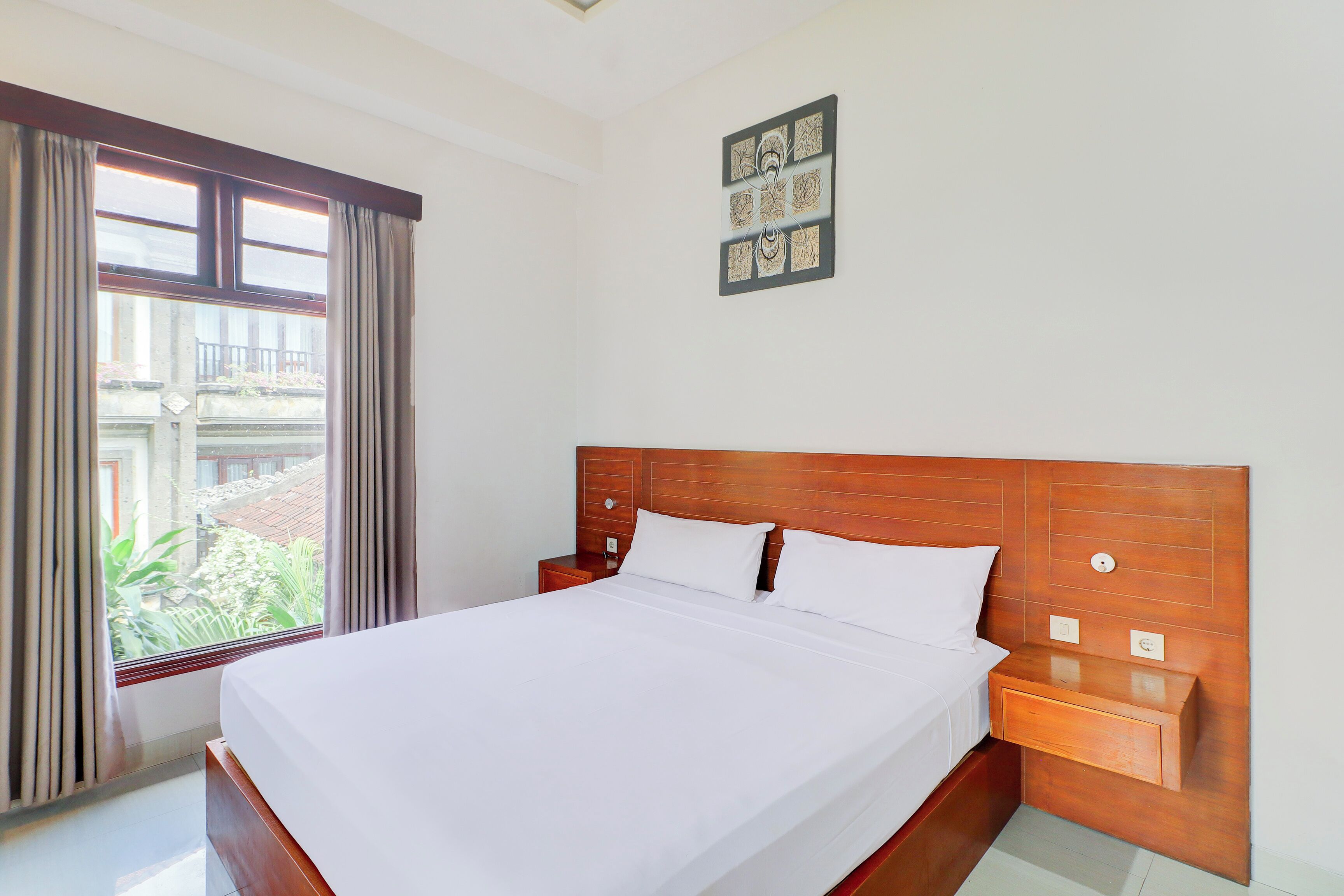 A neatly made bed in a hotel room with a wooden headboard, a bedside table, and a window with curtains.