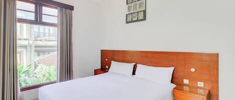 A neatly made bed in a hotel room with a wooden headboard, a bedside table, and a window with curtains.