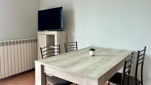 Un comedor con una mesa de madera clara, sillas negras, una planta pequeña en maceta y un televisor sobre un mueble de madera.