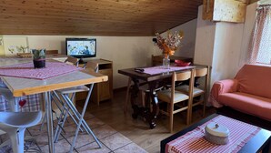 Ein gemütliches Zimmer mit Holzbalkendecke, einem Esstisch mit Stühlen, einem Fernseher, einer Couch und einem kleinen Tisch mit einem dekorativen Gegenstand.