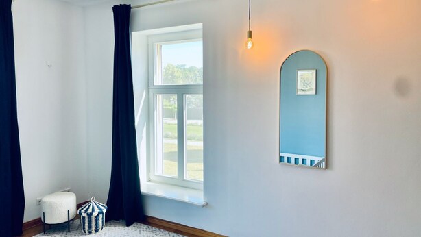Ein Zimmer mit großem Fenster, einem weißen Heizkörper, einem weißen Stuhl, einem blauen Wandspiegel und einem gemusterten Teppich.