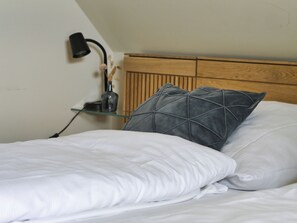 Cama bem arrumada com lençóis brancos e travesseiro cinza, luminária de cabeceira e pequeno vaso com flores secas em um criado-mudo.