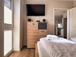 A bedroom with a bed, a wooden wardrobe, a television mounted on the wall, and a window with blinds.