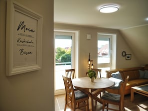 Uma área de refeições com uma mesa redonda, cadeiras e um vaso com flores. Há uma placa emoldurada na parede e uma janela com vista para árvores.