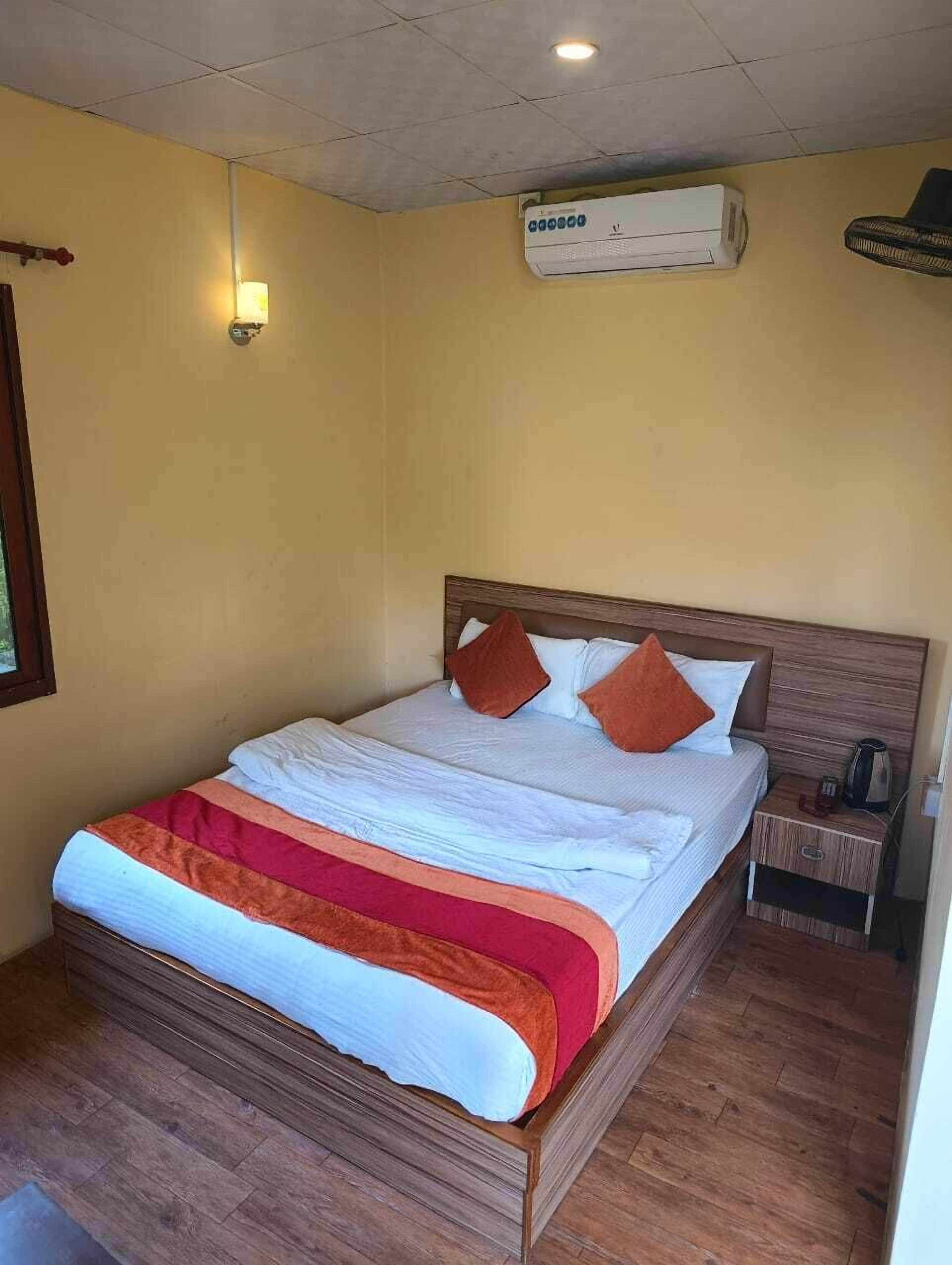 A hotel room with a wooden bed, white bedding, a red and orange blanket, a nightstand with a kettle, and an air conditioning unit on the wall.