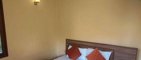 A hotel room with a wooden bed, white bedding, a red and orange blanket, a nightstand with a kettle, and an air conditioning unit on the wall.