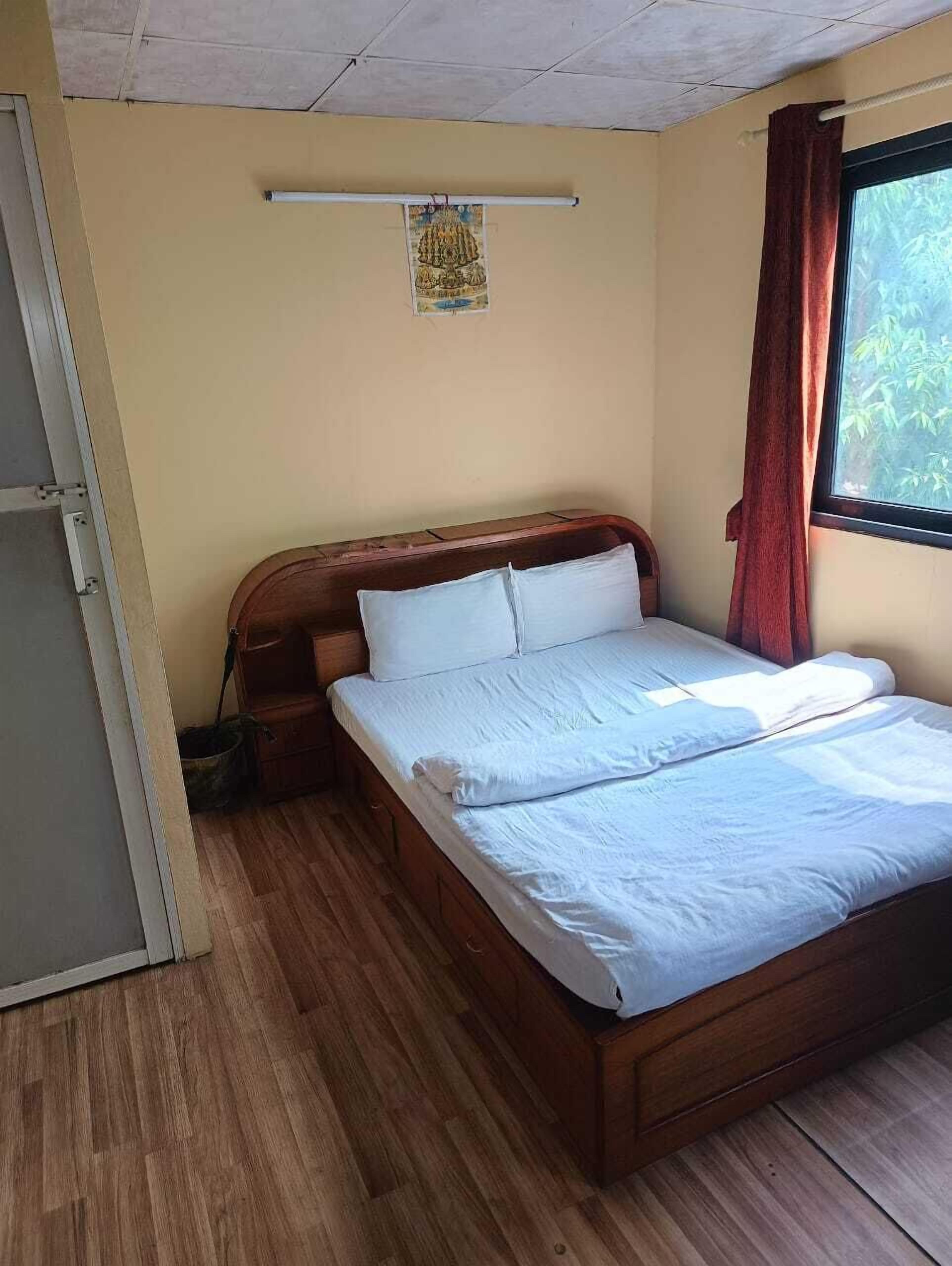Una cama individual con ropa de cama blanca en una habitación con suelo de madera, una ventana con cortinas rojas y una lámpara fijada a la pared.