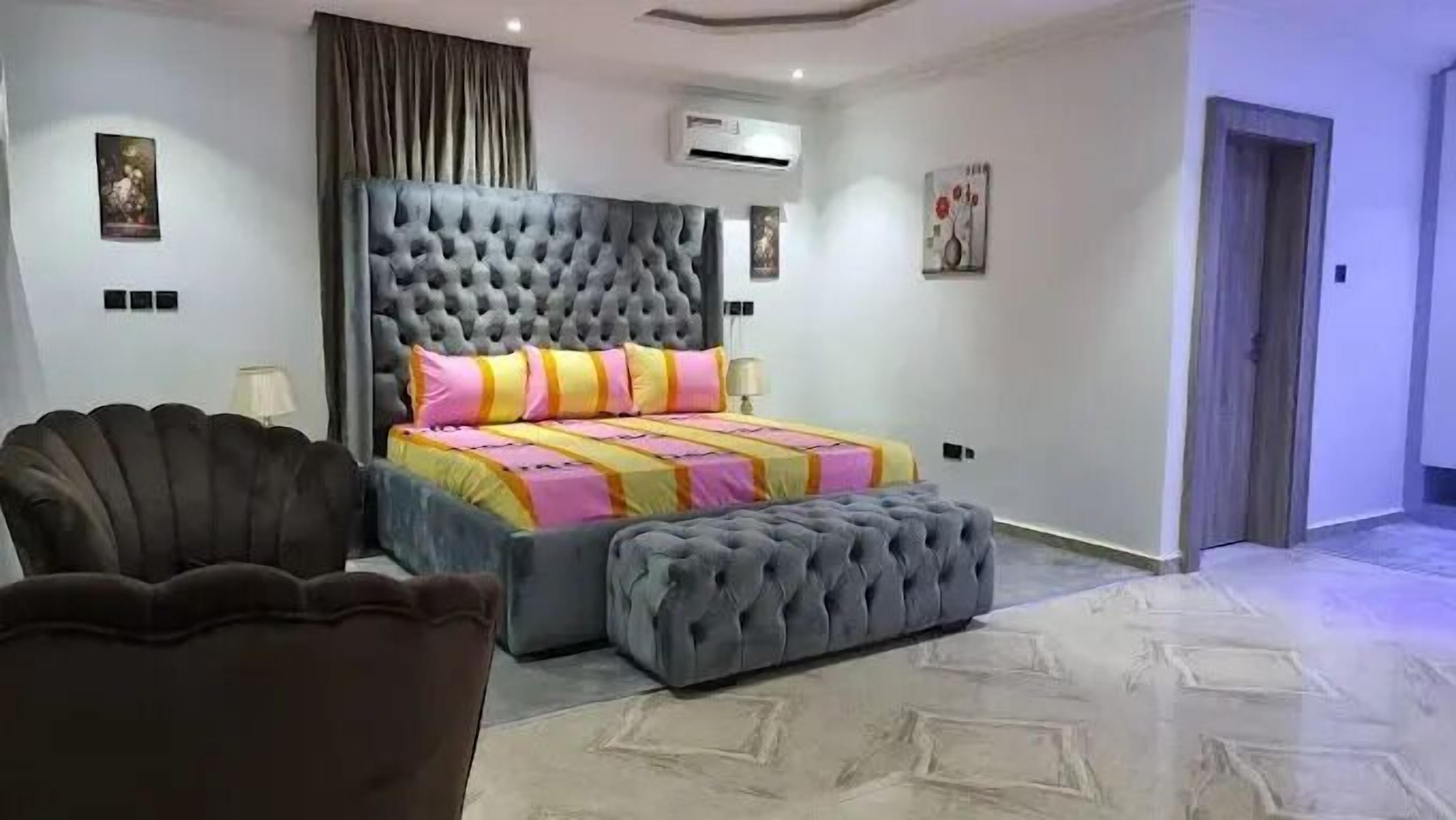 Un dormitorio con cabecera capitonada, cama con ropa de cama colorida, un taburete gris, un sillón marrón y un equipo de aire acondicionado en la pared.
