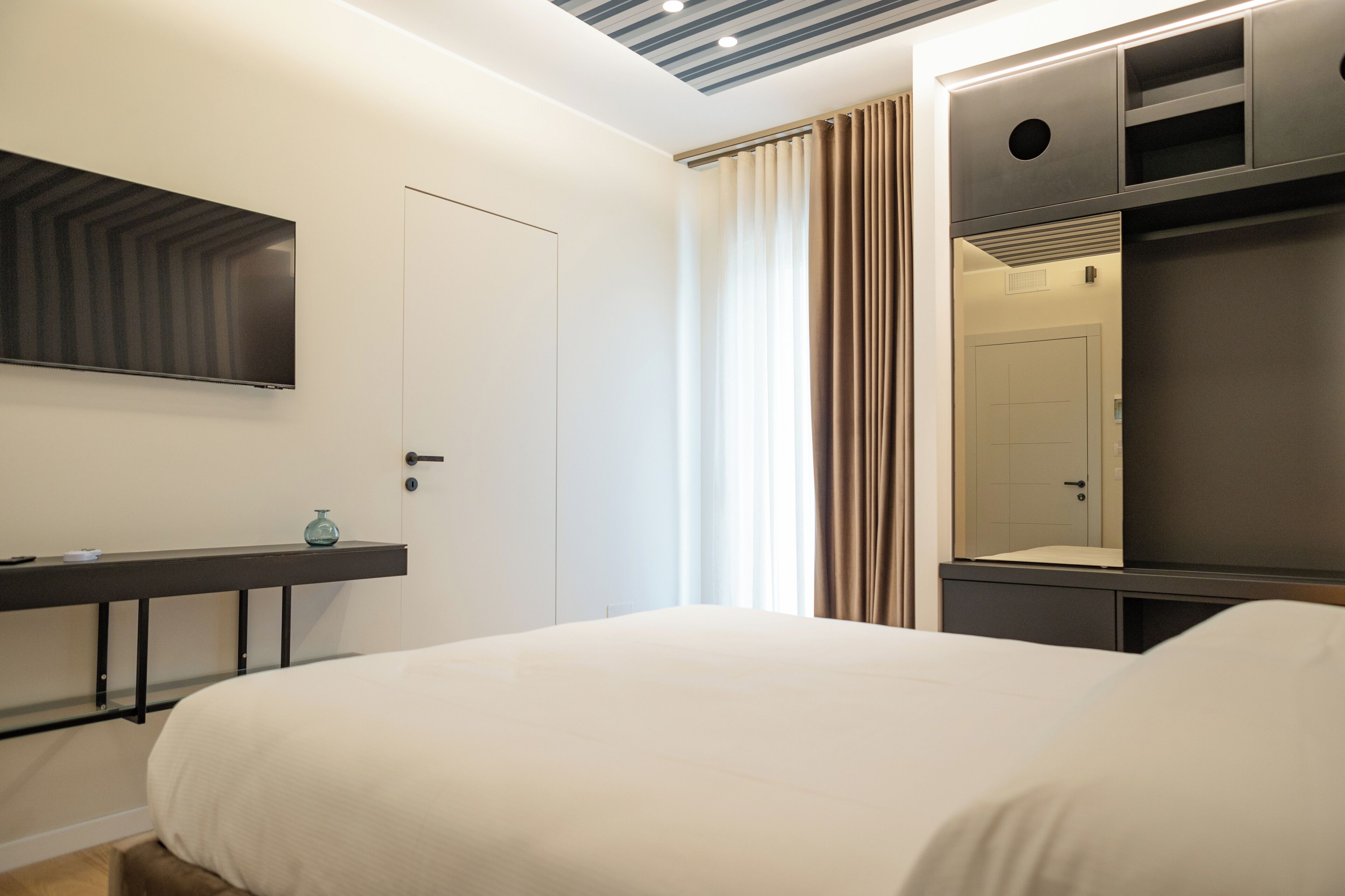 Quarto de hotel moderno com uma cama grande, uma televisão de tela plana na parede, uma mesa de cabeceira com um recipiente de vidro e um guarda-roupa com espelho.