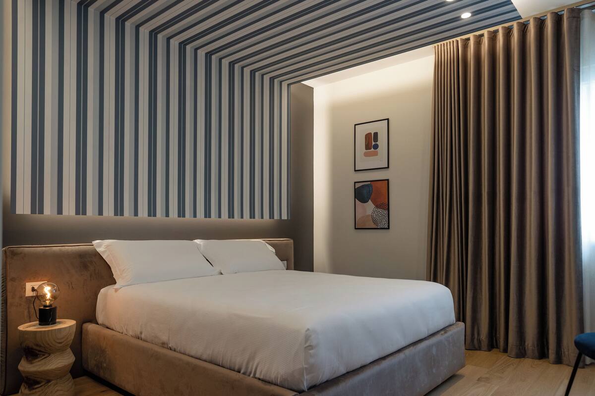 Une chambre moderne avec un grand lit, une table de chevet avec une lampe, un papier peint à rayures et une vue sur l’extérieur à travers des rideaux.