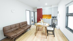A modern living room with a brown leather sofa, a wooden dining table with two chairs, and a kitchen area with white cabinets and a red accent wall. - Melbourne Studio Apartments Hotel @ 138 Elgin Carlton (Carlton)