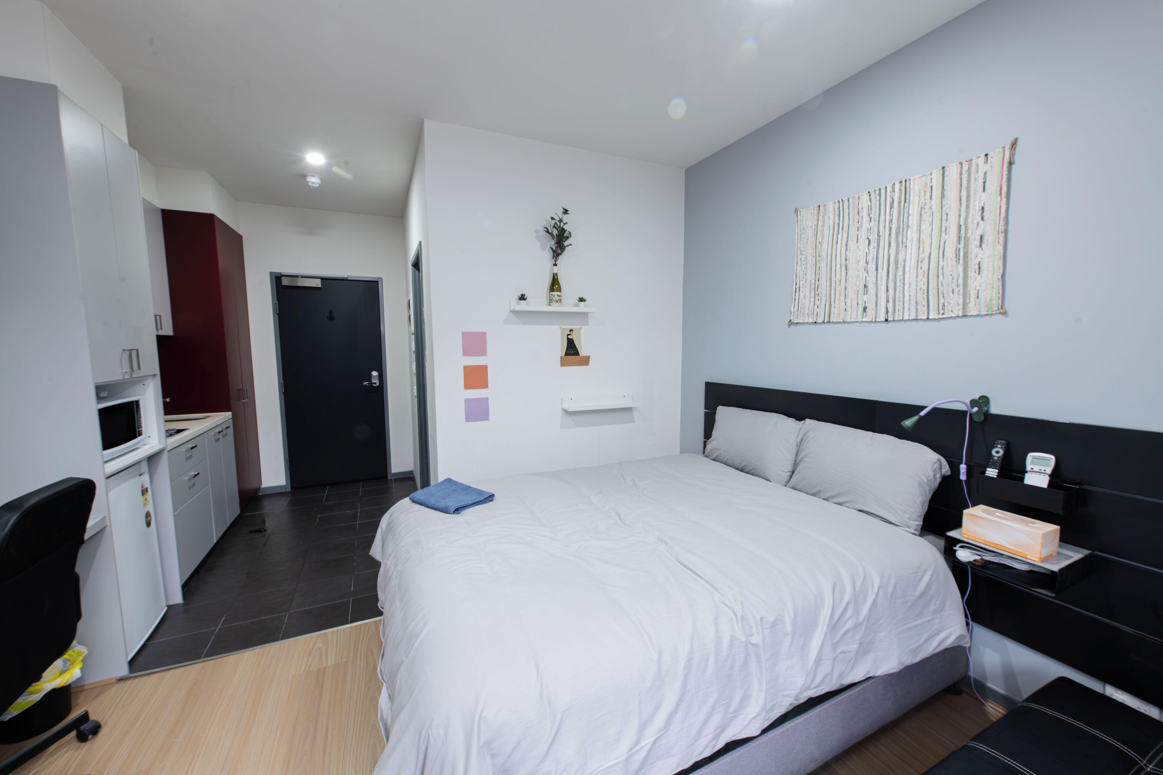 A modern hotel room with a large bed, a desk, and a chair. There is a door leading to the bathroom and a small shelf with decorative items.