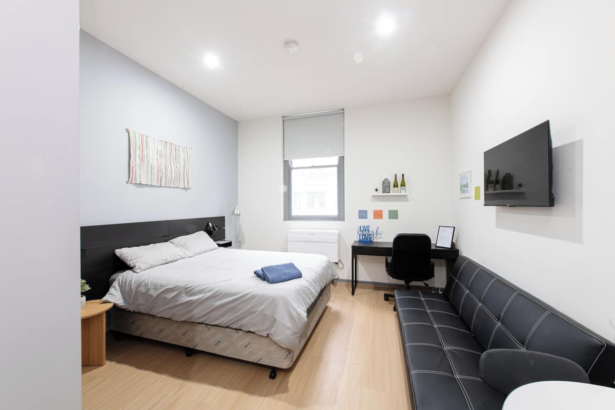 A bedroom with a bed, a desk, a chair, a sofa, a window, and a flat-screen TV on the wall.