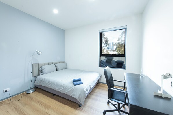 Ein Schlafzimmer mit Bett, Schreibtisch, Stuhl, Lampe und einem Fenster mit Blick auf Gebäude.