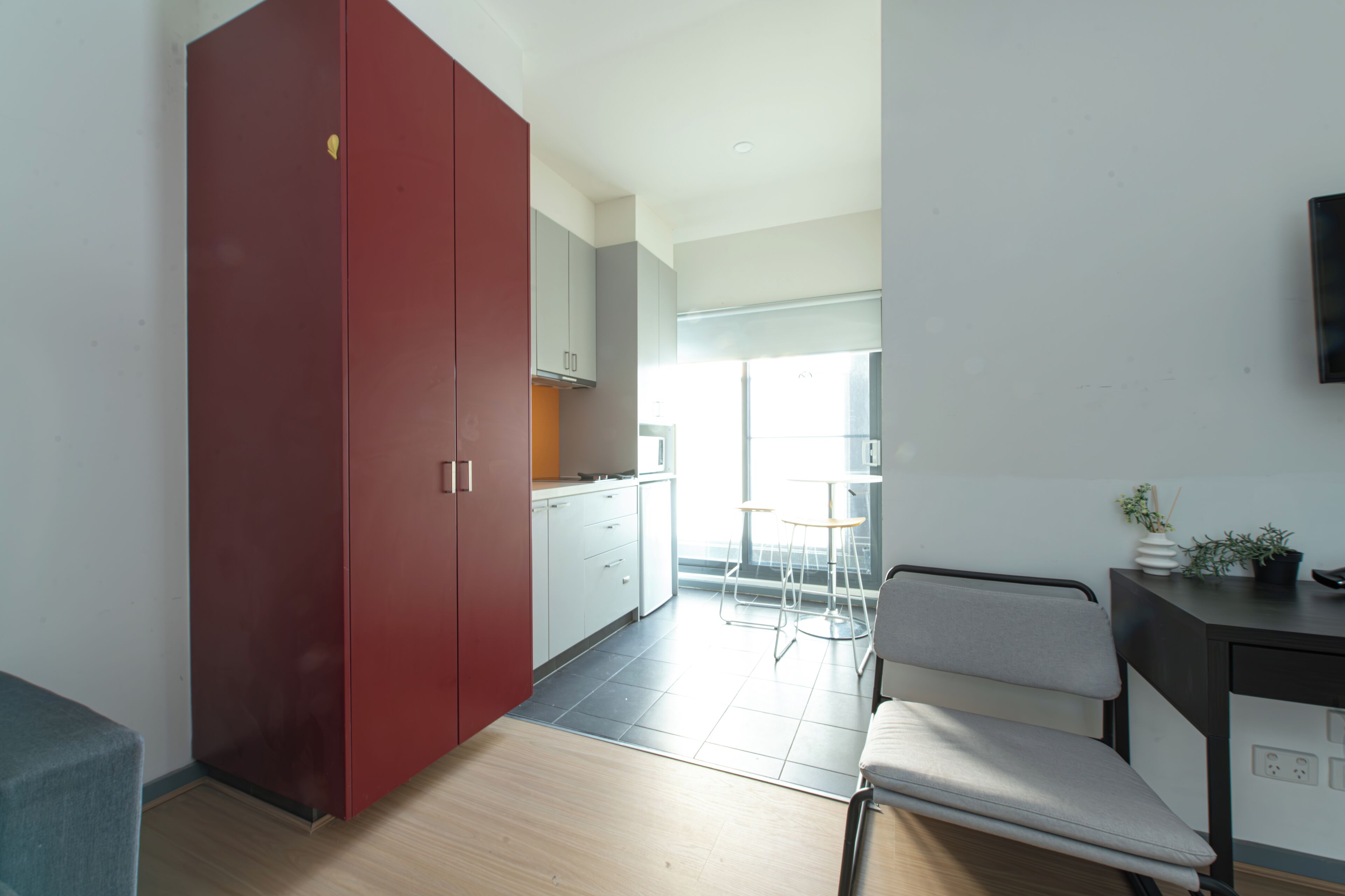 A modern kitchen with a red wardrobe, a dining area with a grey chair, and a small table.