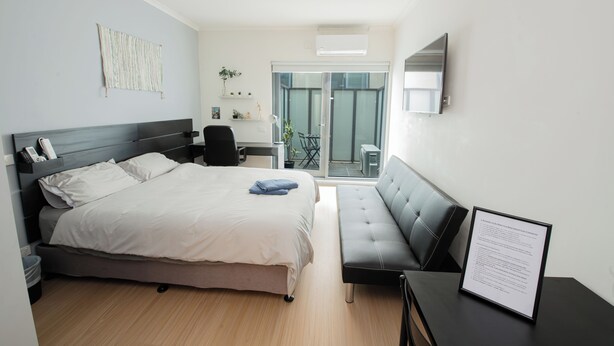 A modern hotel room with a bed, a sofa, a desk, and a balcony with a view. - Melbourne Studio Apartments Hotel @ 138 Elgin Carlton (Carlton)