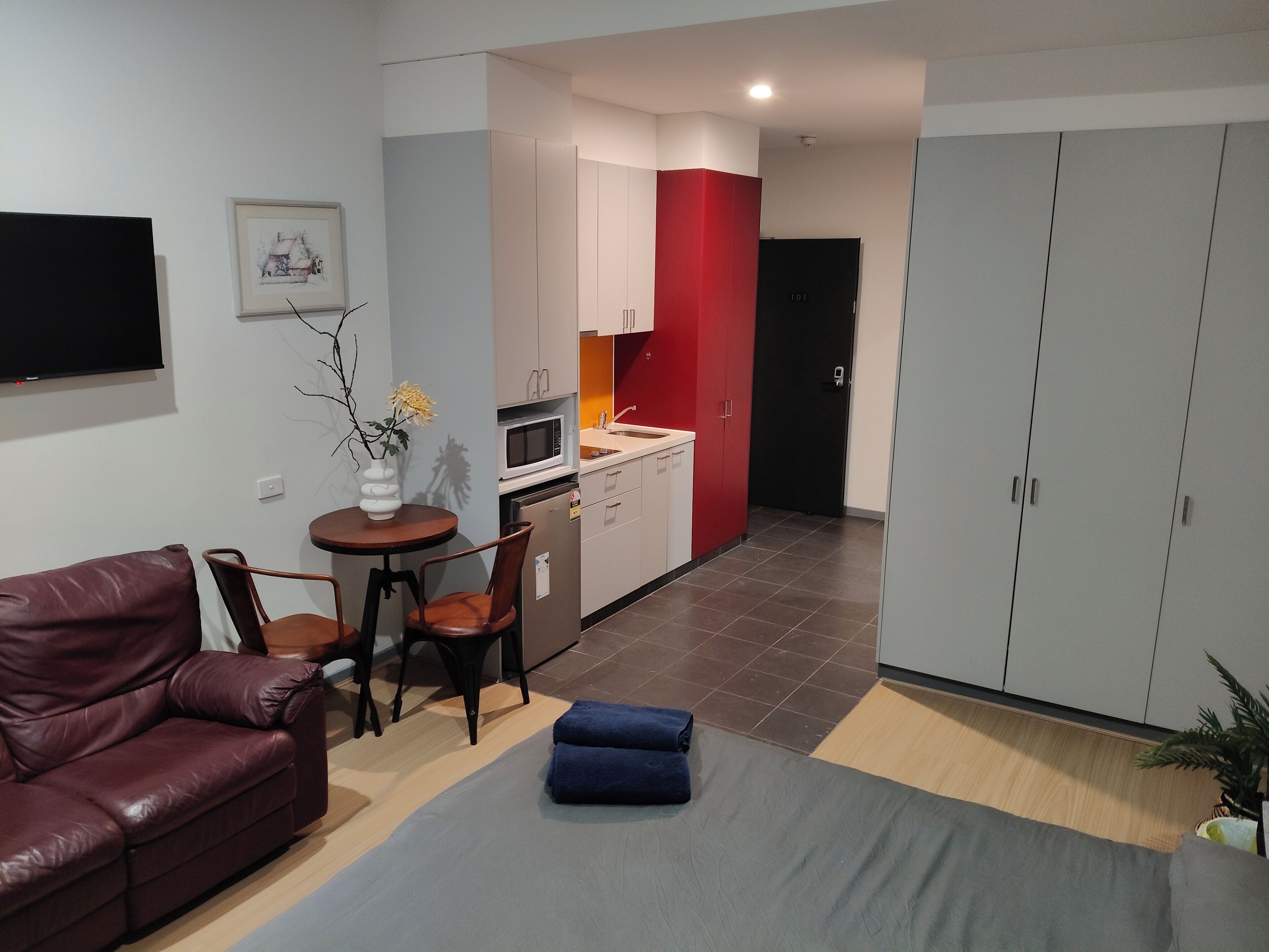 Uma sala de estar moderna com um sofá de couro, uma mesa redonda pequena e um banquinho azul. Uma cozinha com armários brancos e uma parede de destaque vermelha é visível ao fundo.