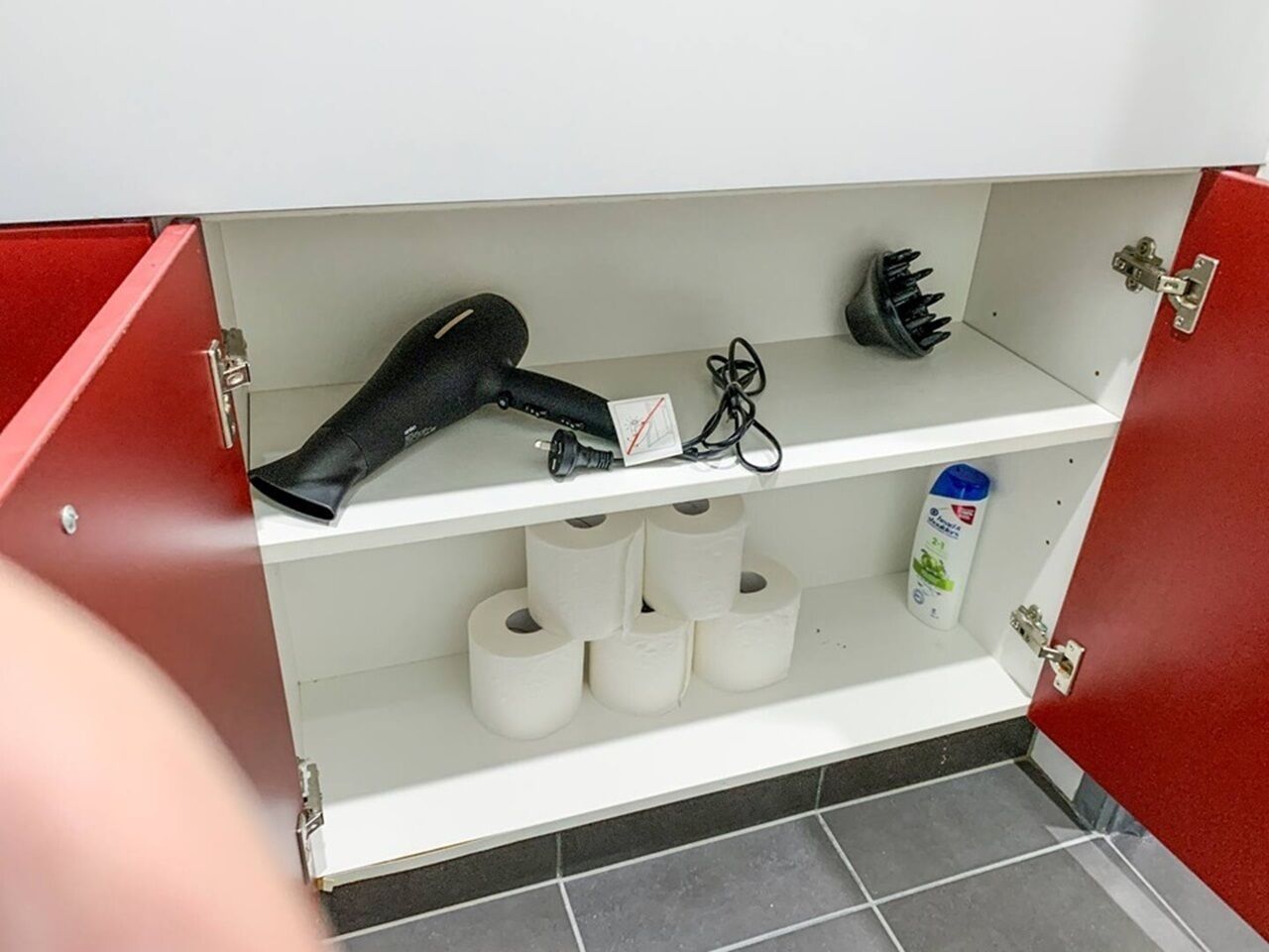Un baño con un armario rojo que contiene un secador de pelo, un cepillo negro para el cabello y una botella de jabón para manos. Hay varios rollos de papel higiénico en el estante inferior.