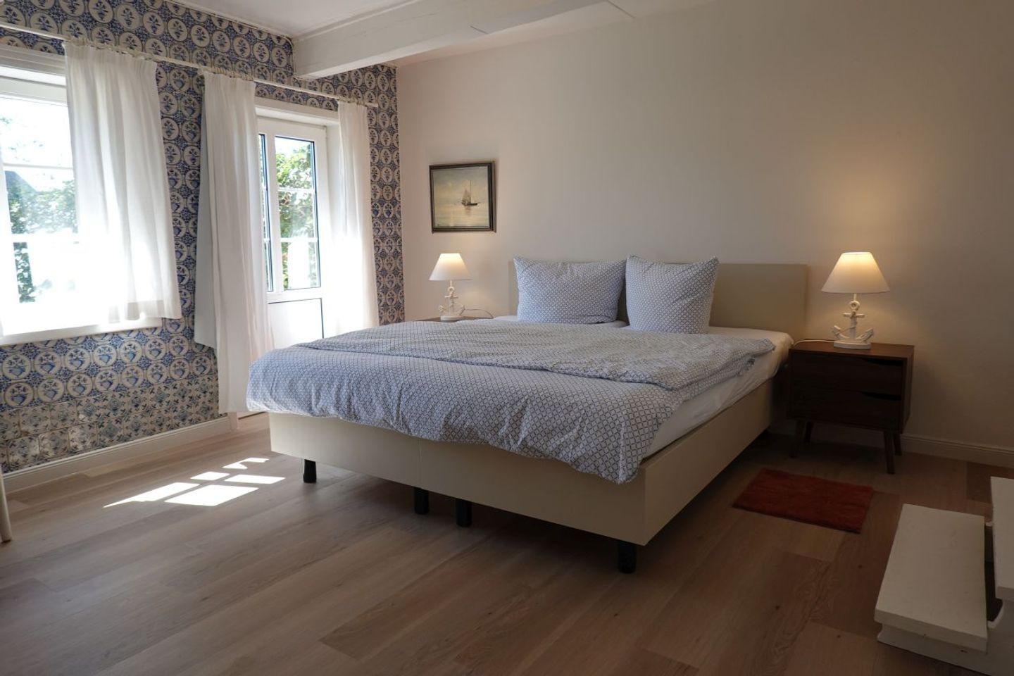 Une chambre à coucher avec un lit, deux tables de chevet, une fenêtre avec des rideaux et un tableau encadré au mur.