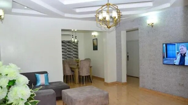 A modern living room with a grey sofa, a small ottoman, a dining area with a table and chairs, and a TV mounted on the wall. - Sterling Lakeview Apartment  (Port Harcourt)